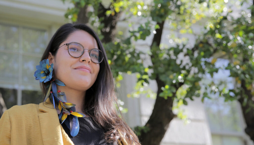 Photo of Astolfi standing in front of a soapbark tree on the UC Berkeley campus, wearing an earring of macaw feathers.