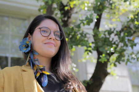 Photo of Astolfi standing in front of a soapbark tree on the UC Berkeley campus, wearing an earring of macaw feathers.