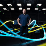 Michael Yartsev stands in a darkened room with his hands on his hips. Yellow and teal lights swirl around him.