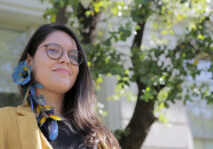 Photo of Astolfi standing in front of a soapbark tree on the UC Berkeley campus, wearing an earring of macaw feathers.