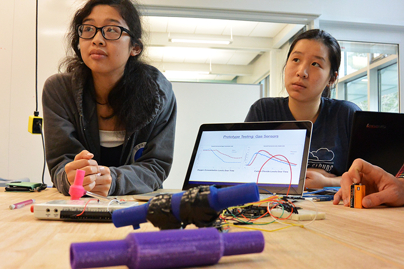 candid photo of two students at workbench listening to lecture with laptop and testing prototypes in front of them